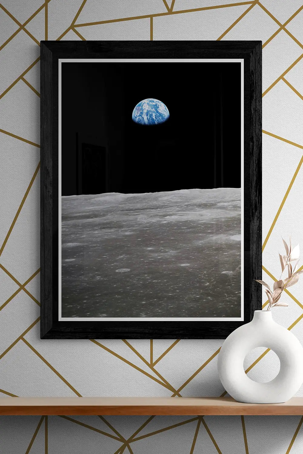 Framed Apollo 11 Earthrise poster showcasing the iconic view of Earth rising above the Moon’s surface, styled on a wooden shelf with a modern vase and geometric wallpaper backdrop.