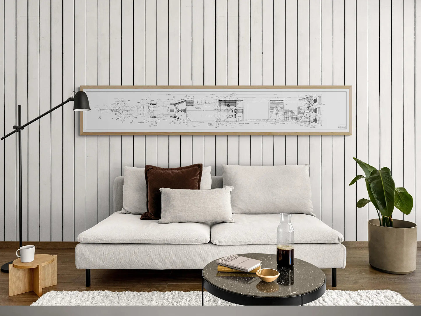 Apollo Saturn V | Rocket Blueprint Posters | Technical Diagram | NASA | A modern living room featuring a light gray sofa with brown and beige pillows, a black floor lamp, and a small wooden side table. The wall above the sofa showcases a light wood-framed blueprint of the NASA Saturn V rocket, with black and white technical drawings on a white paneled background.