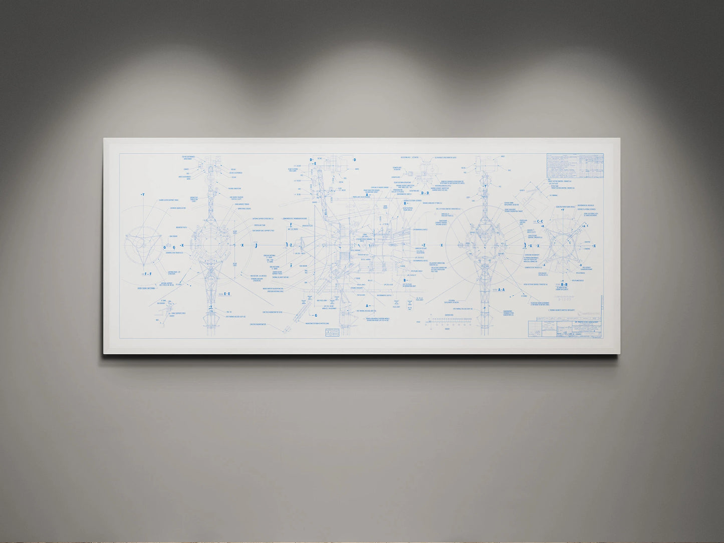 Voyager Space Probes Blueprint | Rocket Blueprint Posters | NASA | JPL | A white-framed blueprint of the NASA Voyager Probes is displayed on a gray wall. The blueprint, featuring a white background with blue annotations, showcases detailed schematics and labels of the spacecraft. Overhead lighting highlights the intricate design, emphasizing the technical precision of the drawings.