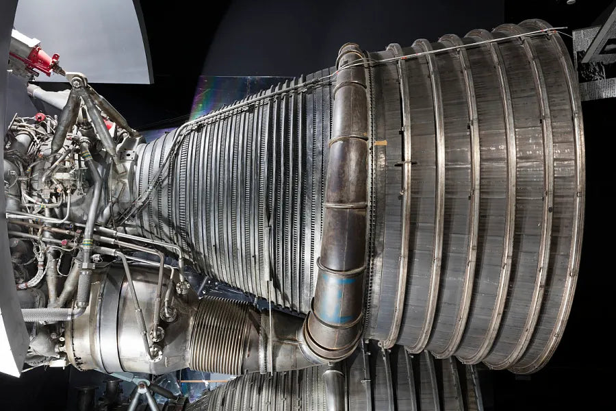 Side view of an F1 rocket engine showing the ribbed nozzle and intricate network of pipes and components. The metallic textures and complex engineering are highlighted against a dark background.