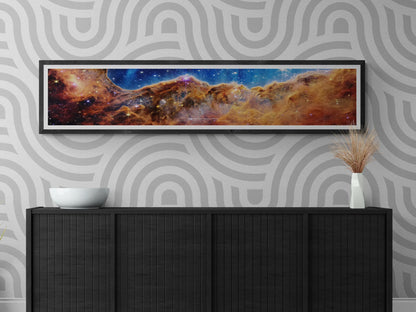 Framed James Webb Space Telescope 'Cosmic Cliffs' panoramic poster displayed in a modern interior, showcasing the vibrant Carina Nebula star-forming region. Ideal space-themed wall art for home or office décor.