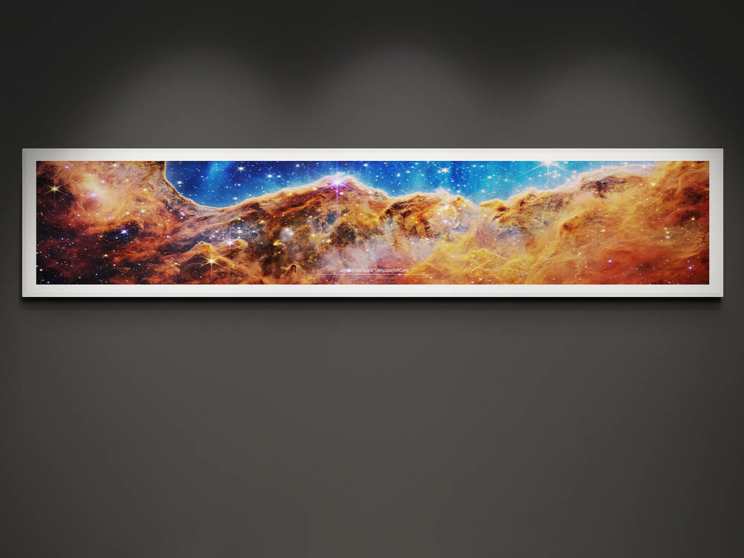 Framed panoramic poster of the James Webb Space Telescope's 'Cosmic Cliffs' image, showcasing a vibrant star-forming region in the Carina Nebula with rich hues of orange, red, and blue, against a dark wall backdrop.