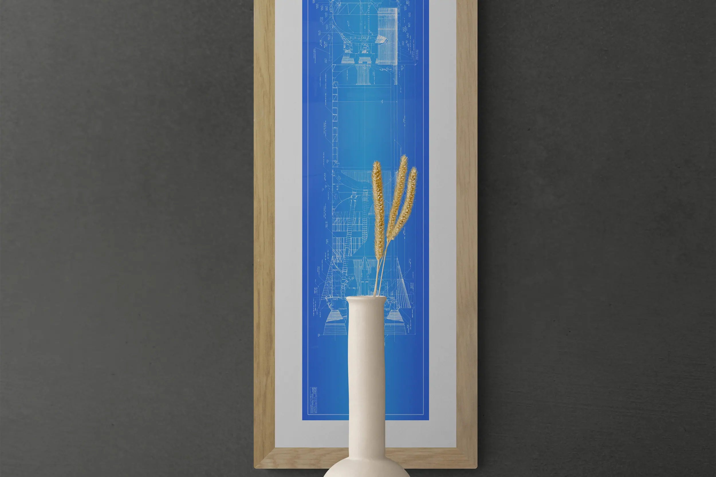 Rocket Posters | NASA | Saturn V | The image features a vertical framed Saturn V rocket blueprint poster with a wooden frame on a dark gray wall. Detailed technical drawings in blue are visible. A white vase with dried wheat stalks is positioned at the bottom center of the poster.