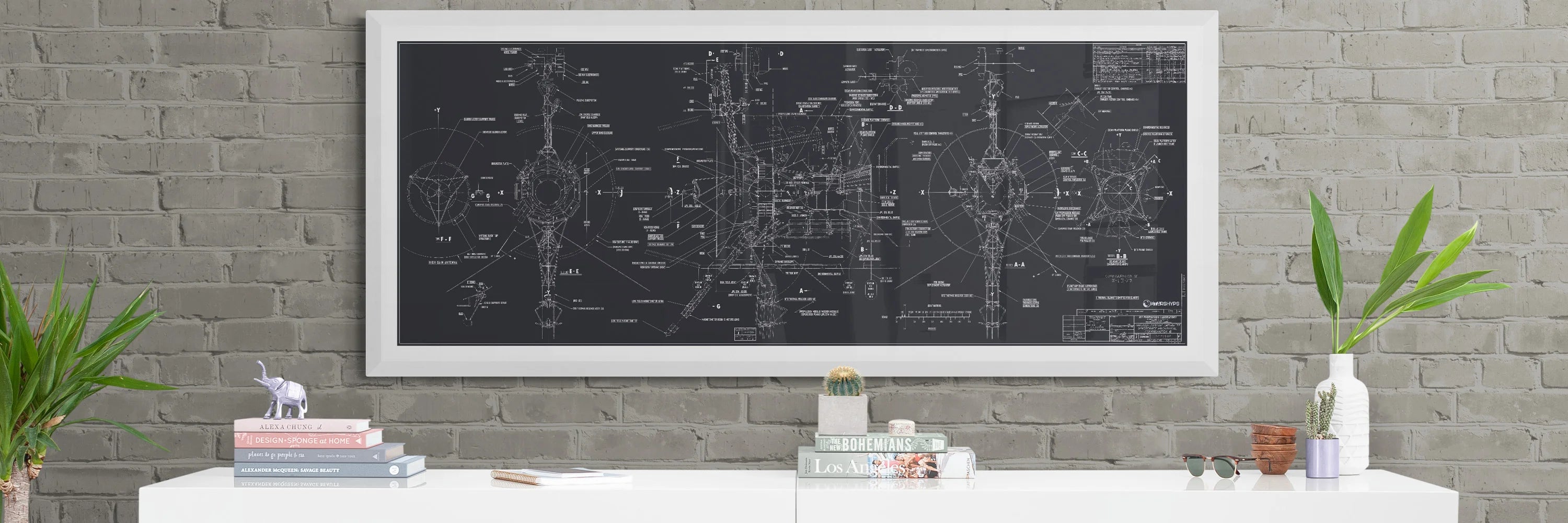 Voyager Space Probes Blueprint | Rocket Blueprint Posters | NASA | JPL | The framed Voyager probes blueprint poster, filled with intricate technical drawings and annotations, is mounted on a gray brick wall. Below, a white cabinet is styled with a mix of books, a potted plant, and decorative items.