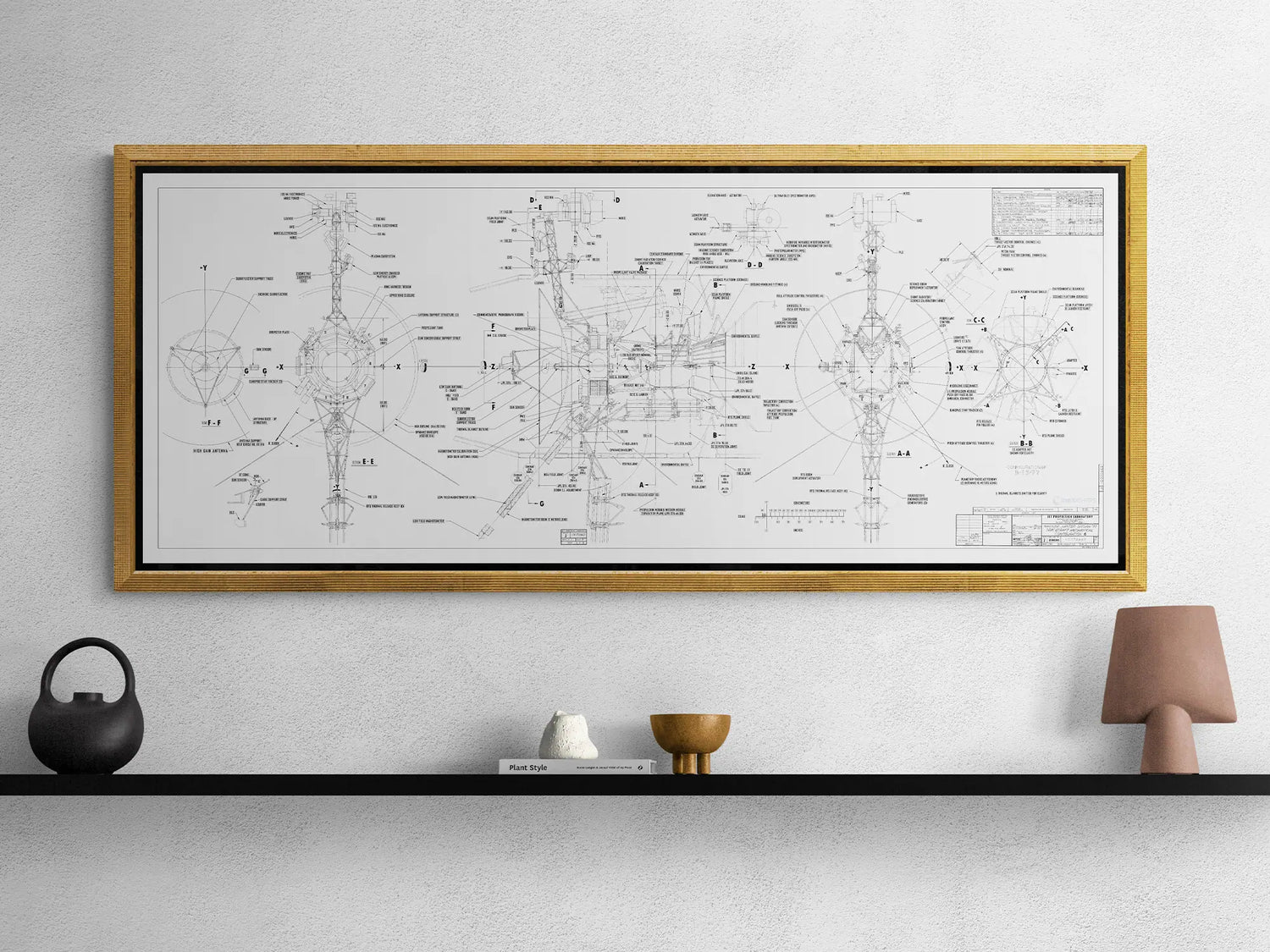 Voyager Space Probes Blueprint | Rocket Blueprint Posters | NASA | JPL | A framed technical drawing of the Voyager probes hangs on a white wall. The drawing, encased in a gold frame with a black border, features detailed schematics and labels of various components of the spacecraft. Below the frame, a minimalist black shelf holds a few small decorative items, including a black teapot, a white ceramic piece, and a small brown lamp.